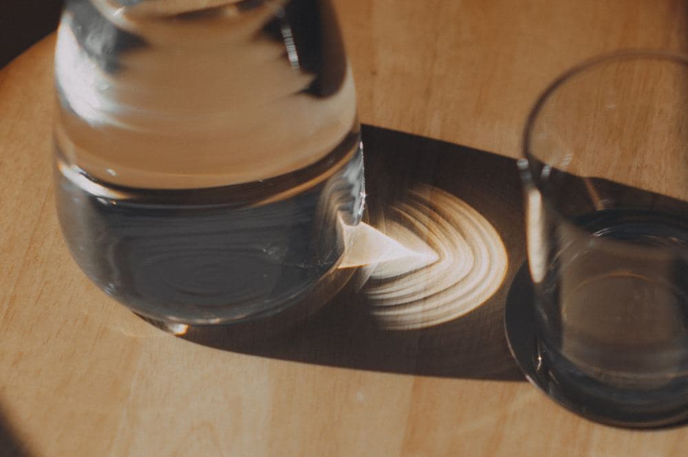 Glaskaraffe und Wasserglas auf einem Holztisch in der Praxis, das Sonnenlicht wirft einen weichen Schatten